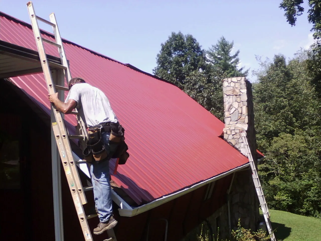 Roofer on a ladder beside a red metal roof and stone chimney — Soffit & Fascia Repair by Roofing Brothers in Whitesboro